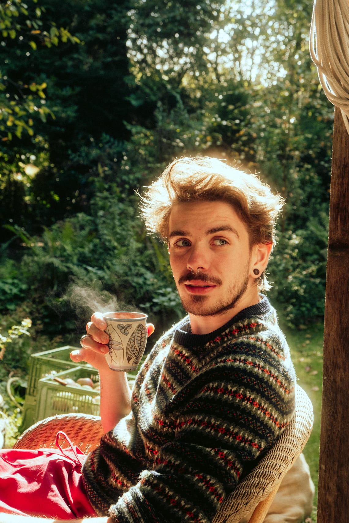       A young man in a knit sweater sitting in a sunny garden, holding a steaming ceramic mug, glancing over his shoulder at the camera.